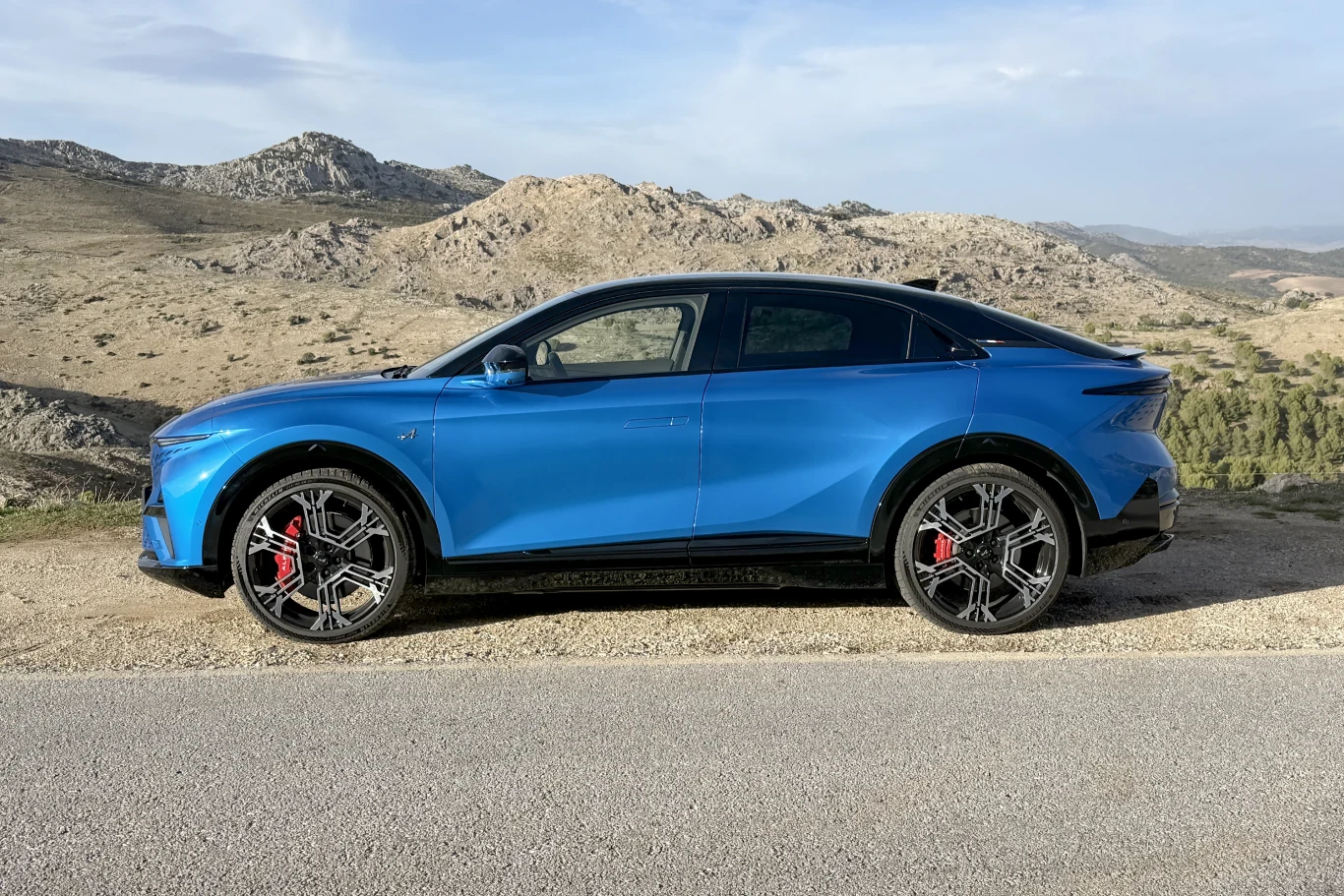 Niebieski samochód sportowy klasy SUV zaparkowany przy asfaltowej drodze na tle górzystego, kamienistego krajobrazu. Pojazd wyróżniają duże, czarne felgi z czerwonymi zaciskami hamulcowymi.