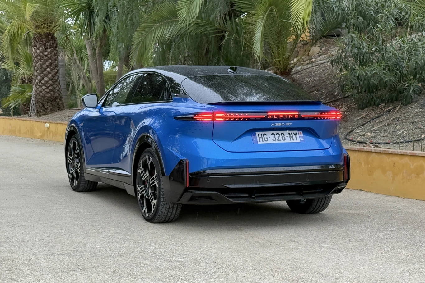 Niebieski nowoczesny SUV Alpine parkuje na asfaltowej drodze w otoczeniu tropikalnej roślinności, auto wyróżnia się charakterystycznym kształtem świateł LED oraz sportowym wykończeniem tylnej części nadwozia.