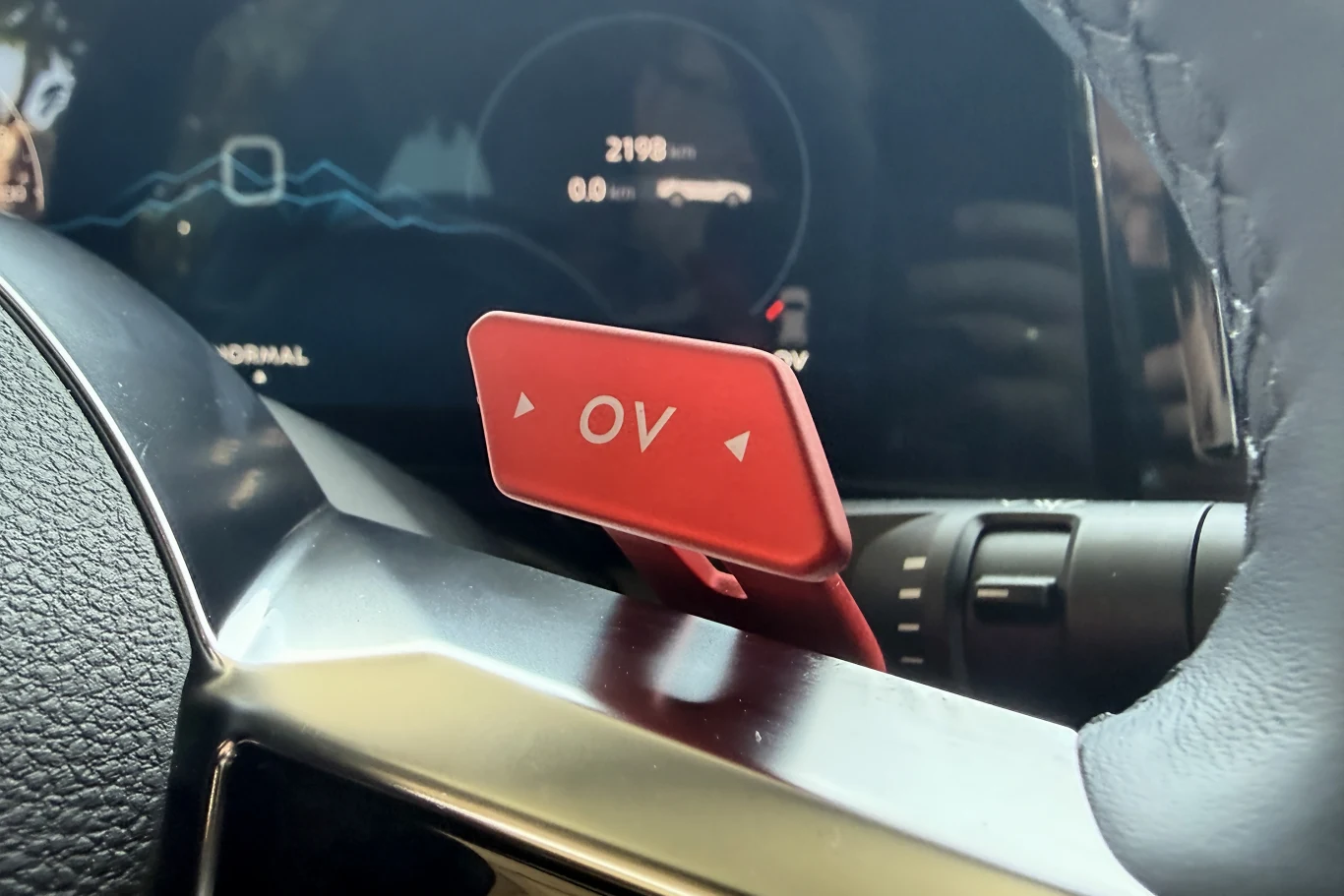 Czerwony przycisk funkcyjny z napisem OV umieszczony przy kierownicy samochodu, w tle widoczny cyfrowy wyświetlacz deski rozdzielczej z informacjami na temat jazdy.