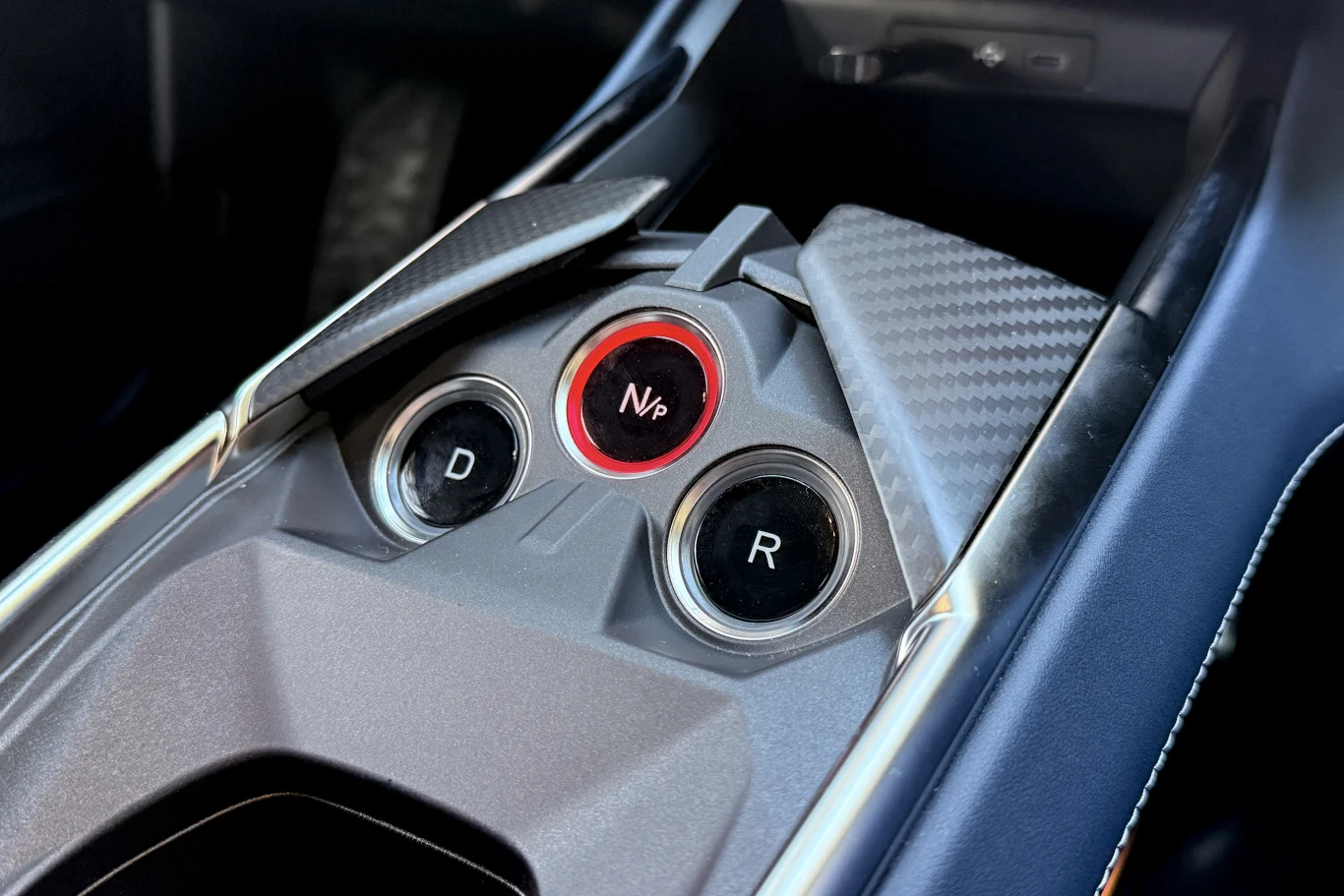 Centralna część konsoli samochodowej wyposażona w trzy przyciski do sterowania automatyczną skrzynią biegów oznaczone literami D, N oraz R, otoczona detalami z materiału przypominającego włókno węglowe.