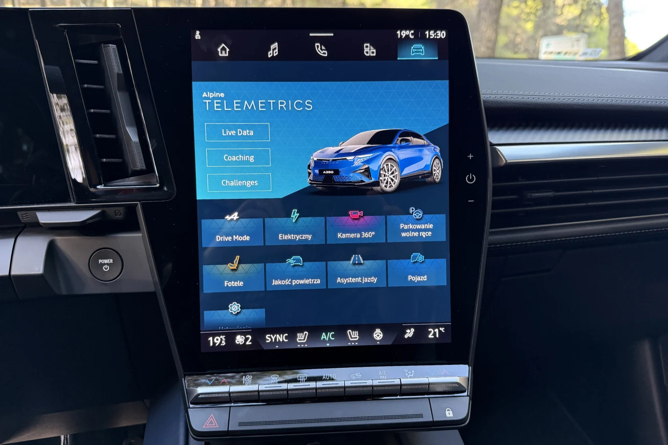 Wyświetlacz multimedialny samochodu prezentujący ekran systemu telemetrii, z widoczną grafiką niebieskiego samochodu, różnymi ikonami i opcjami dotyczącymi parametrów jazdy, jakości powietrza oraz ustawień pojazdu.