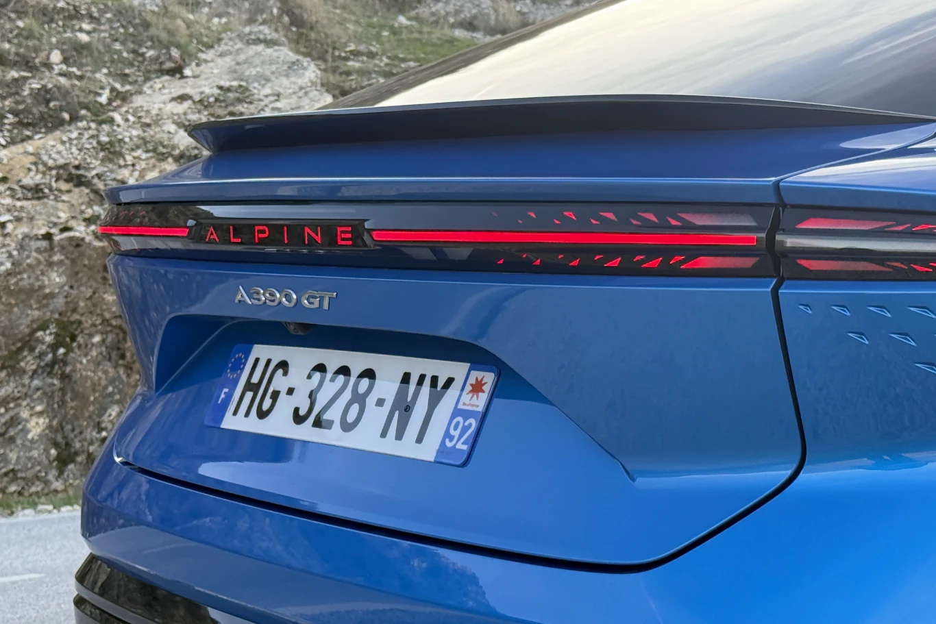 Tylna część niebieskiego samochodu Alpine A290 GT z wyraźnie widocznymi światłami LED, spoilerem oraz francuską tablicą rejestracyjną, otoczona górzystym krajobrazem.