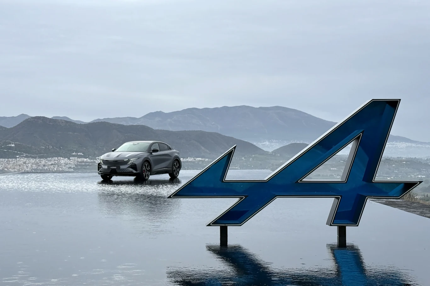 Czarny samochód osobowy stoi na powierzchni odbijającej wodę, na pierwszym planie widoczne jest duże niebieskie logo z literą „A”, w tle góry oraz pochmurne niebo.