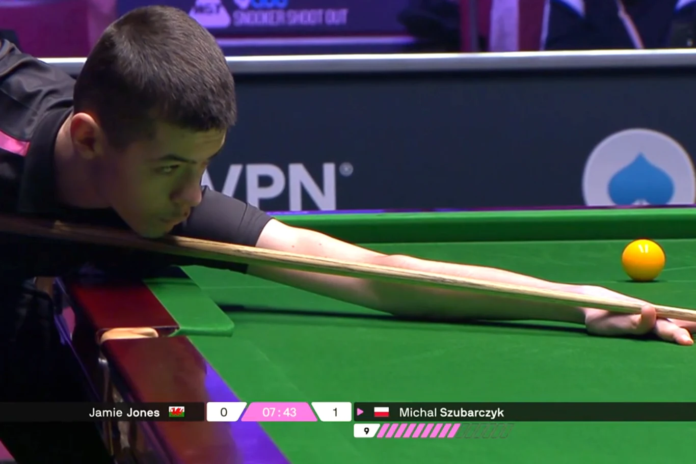 Zawodnik snookera wykonuje precyzyjny strzał, skupiony nad zielonym stołem bilardowym. Obok niego znajduje się żółta bila, widoczne są tablice wyników oraz imię i nazwisko gracza.