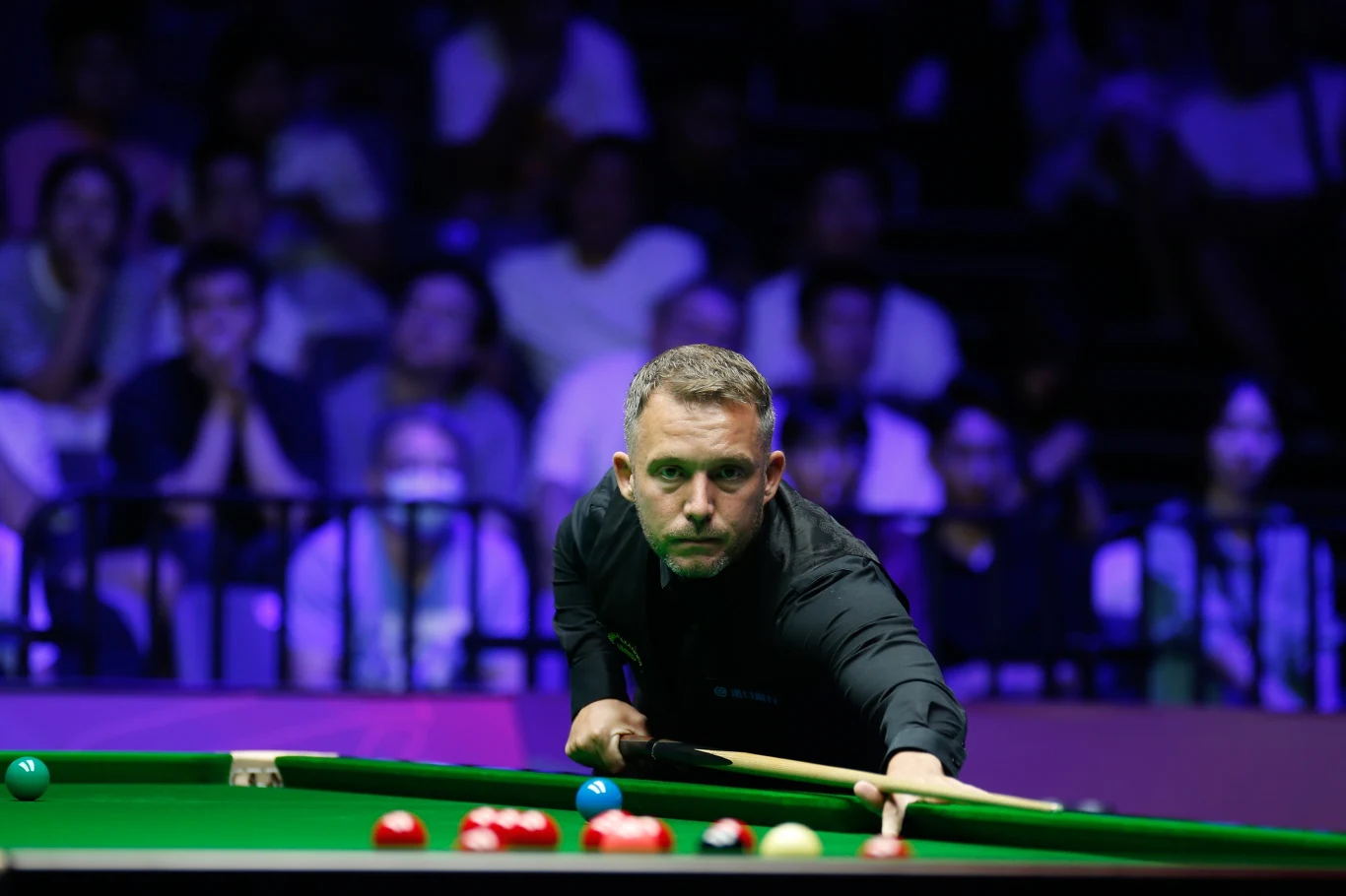 Mężczyzna w czarnej koszuli skupiony nad stołem bilardowym, przygotowuje się do uderzenia kijem snookerowym, na pierwszym planie bile ułożone na zielonym suknie, w tle widownia z rozmytymi sylwetkami kibiców.