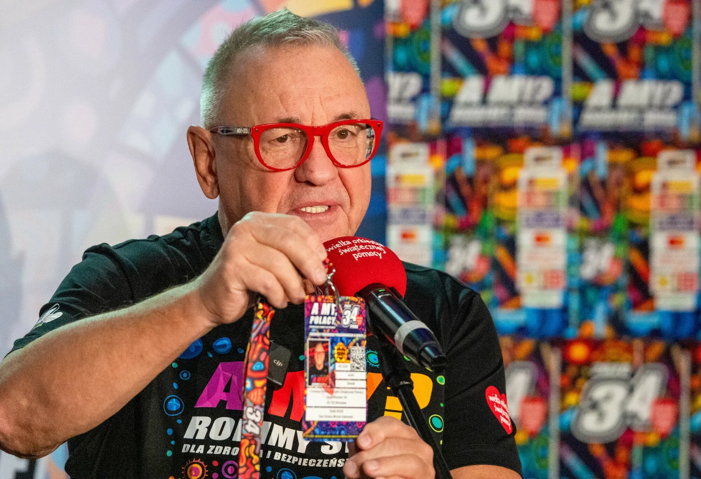 Mężczyzna w okularach o czerwonych oprawkach trzyma w dłoni identyfikator i mówi do mikrofonu, w tle widoczne są kolorowe plakaty promocyjne.