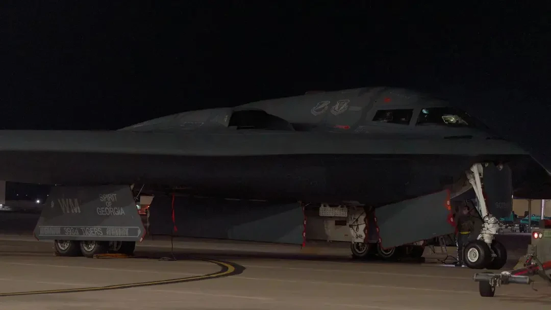 Wojskowy bombowiec stealth B-2 Spirit stojący na pasie lotniska nocą, w otoczeniu oświetlenia technicznego, z widocznymi elementami podwozia i kształtem charakterystycznym dla technologii niewykrywalności radarowej.