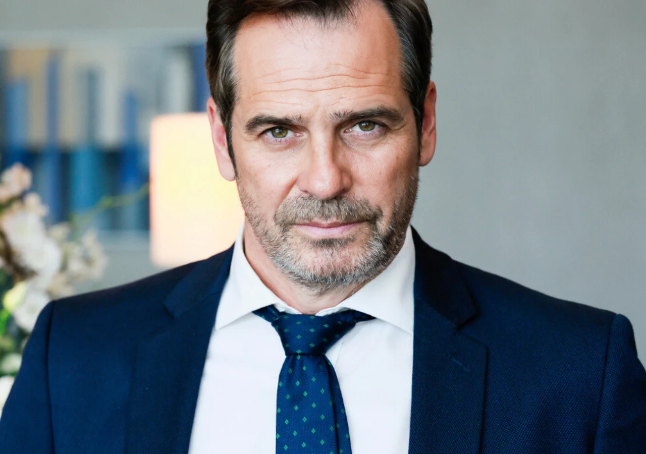 Paweł Deląg Mężczyzna w średnim wieku w granatowym garniturze i krawacie w zielone kropki patrzy pewnie przed siebie, w tle rozmyte elementy wnętrza i roślinność.