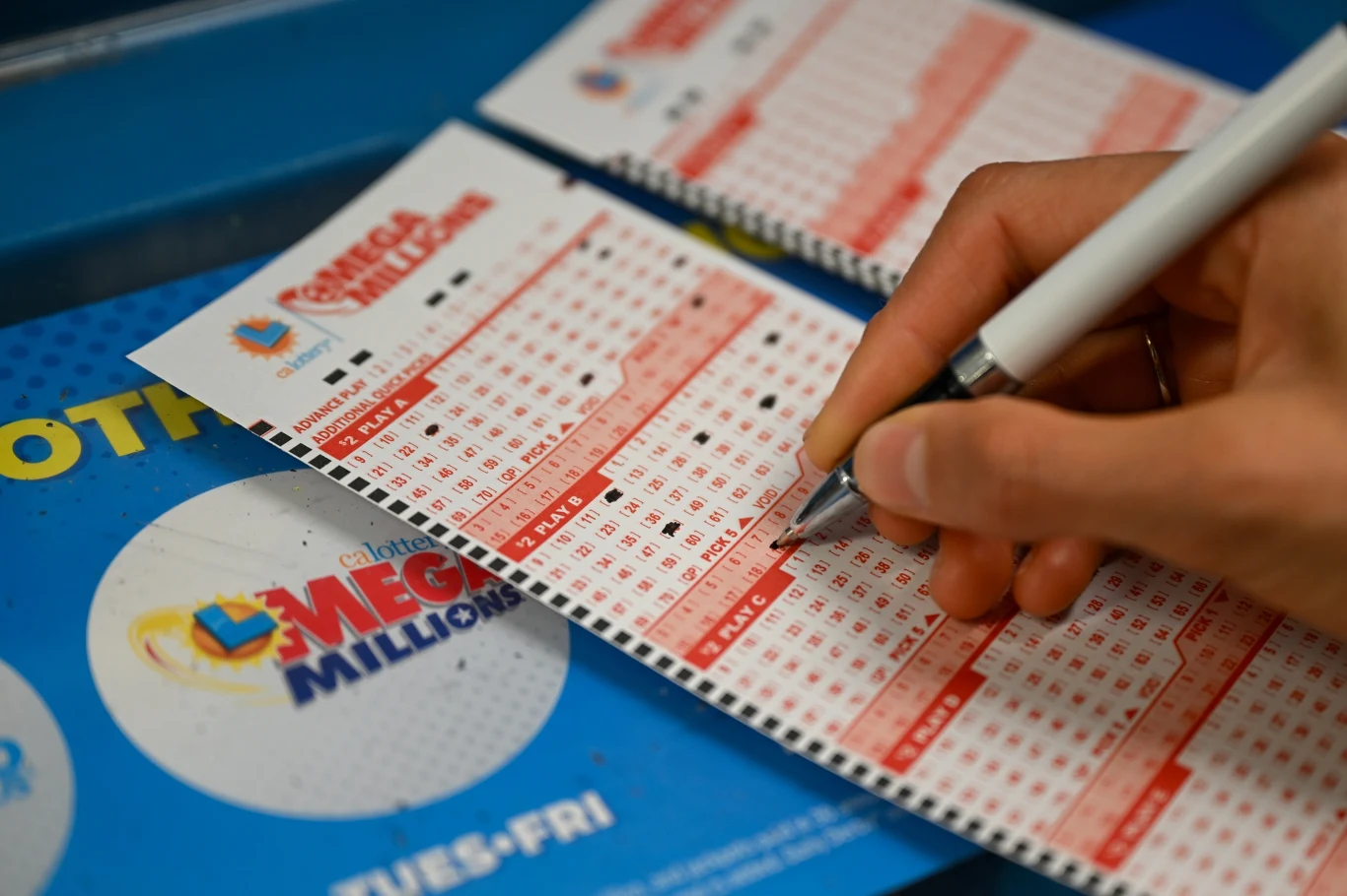 Jackpoty rosną szybciej niż kiedykolwiek (zdj. ilustracyjne) Ręka osoby zaznaczającej liczby na kuponie loterii Mega Millions umieszczonym na niebieskim podkładzie z logo oraz nazwą loterii.