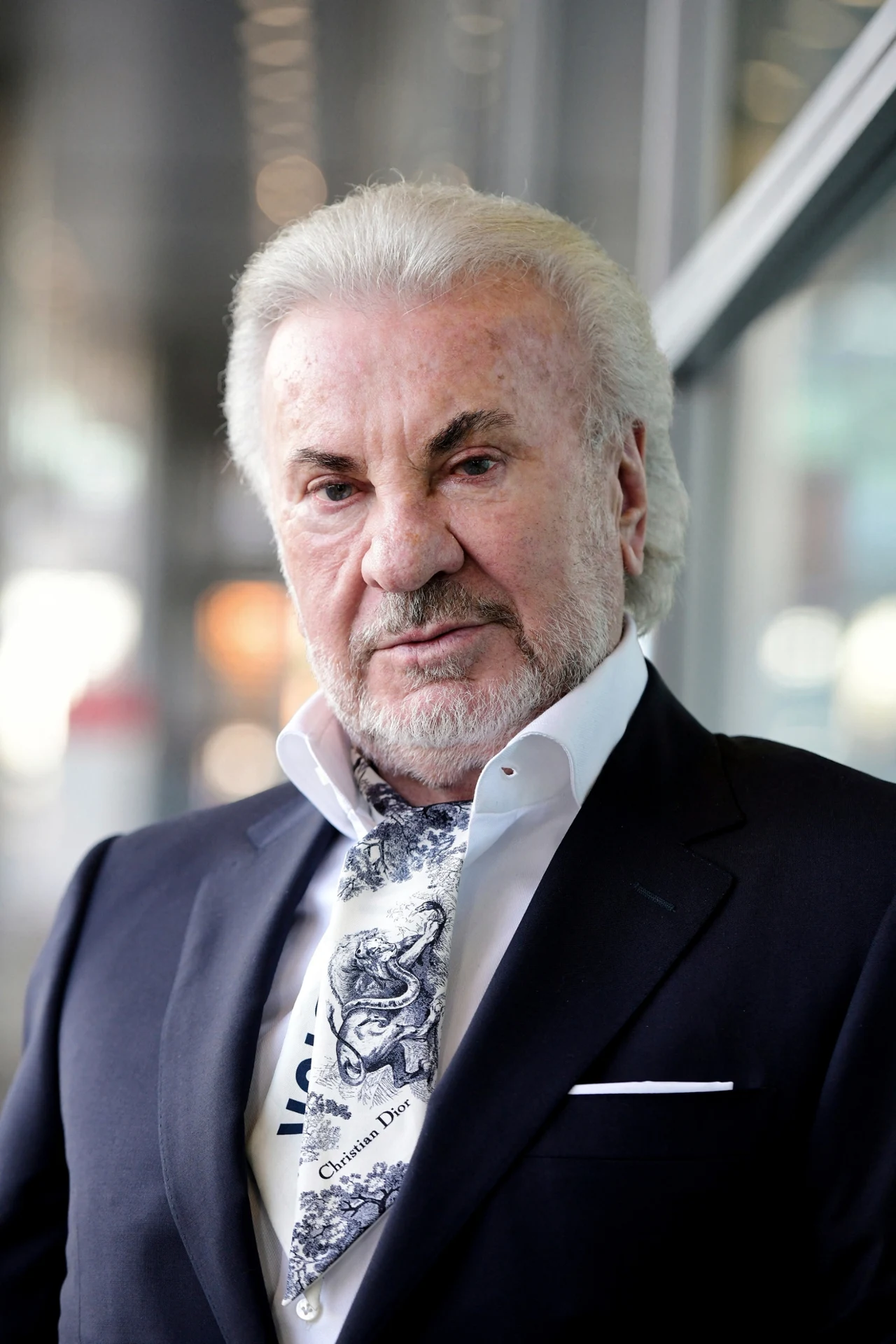 Starszy mężczyzna w eleganckim garniturze z białą poszetką i ozdobnym szalikiem, stojący na tle nowoczesnych przeszklonych ścian, o pewnym wyrazie twarzy i starannie ułożonych siwych włosach.