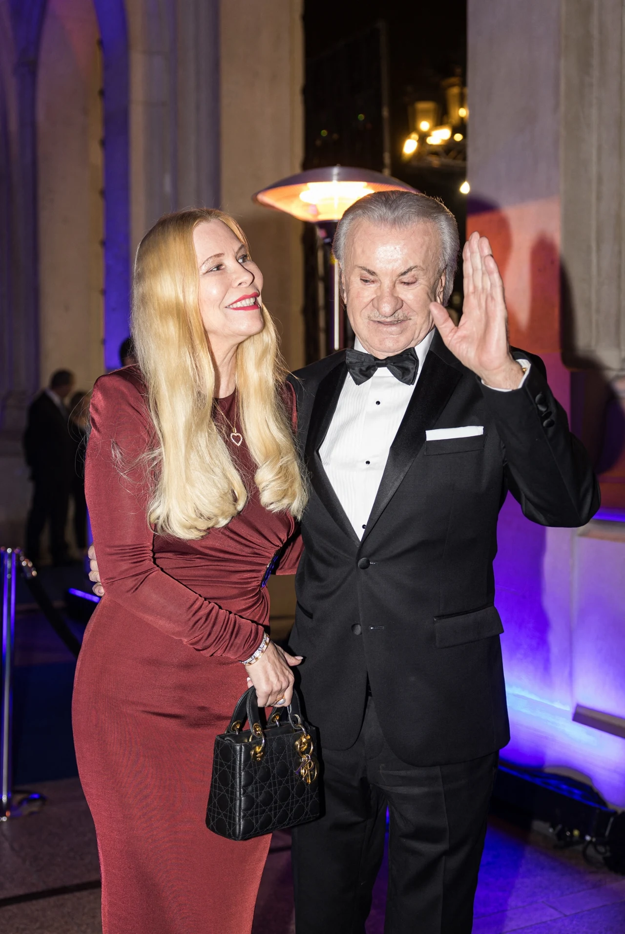 Elegancka kobieta w długiej bordowej sukni i mężczyzna w czarnym smokingu uśmiechają się do siebie podczas oficjalnego wydarzenia wieczorowego, stojąc blisko siebie. Kobieta trzyma czarną torebkę, a mężczyzna unosi dłoń w geście pozdrowienia lub rozmow...