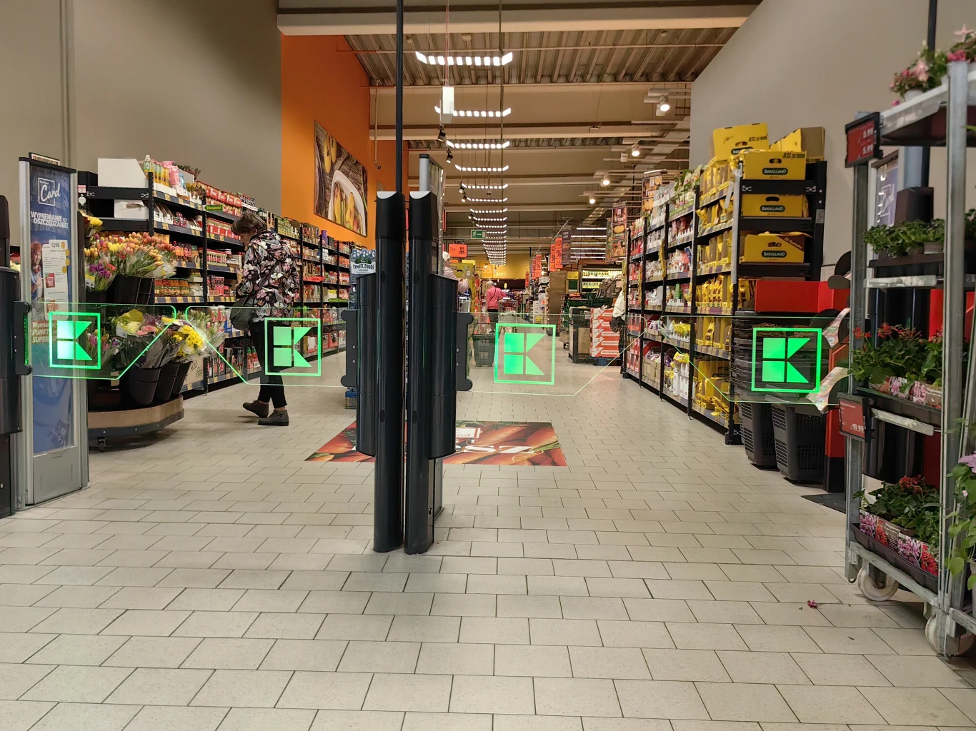 Związkowcy zapowiadają strajk w najbliższą handlową niedzielę, a Kaufland zapewnia, że sklepy będą działać bez przeszkód Alejki supermarketu z wypełnionymi regałami pełnymi produktów spożywczych, w tym owoców, warzyw oraz artykułów pierwszej potrzeby, na pierwszym planie bramki wejściowe zabezpieczające, a w tle widoczni klienci robiący zakupy.