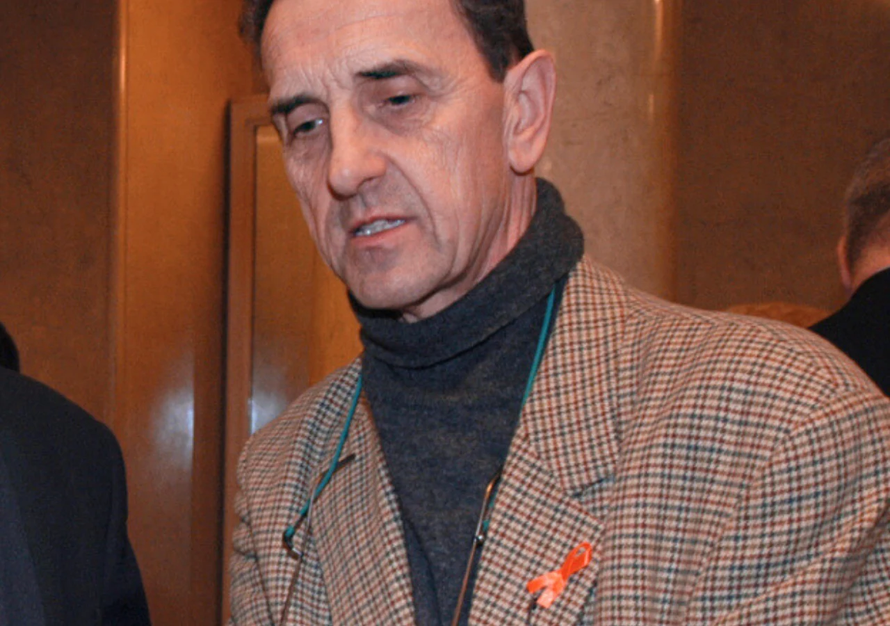 Krzysztof Logan Tomaszewski Mężczyzna w marynarce w kratę i swetrze z golfem, z przypiętą pomarańczową wstążką na klapie, stoi w pomieszczeniu o ciepłym oświetleniu, z widocznym fragmentem innej osoby w tle.