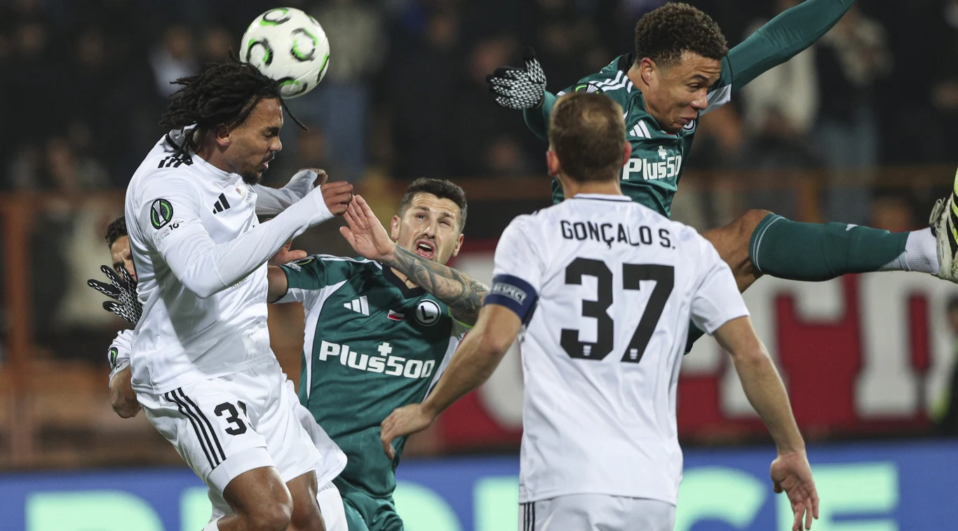 Piłkarze walczą w powietrzu o piłkę podczas meczu, zawodnik w zielonym stroju wyskakuje najwyżej, obok niego znajduje się piłkarz w białym. Kibice i inni zawodnicy w tle.