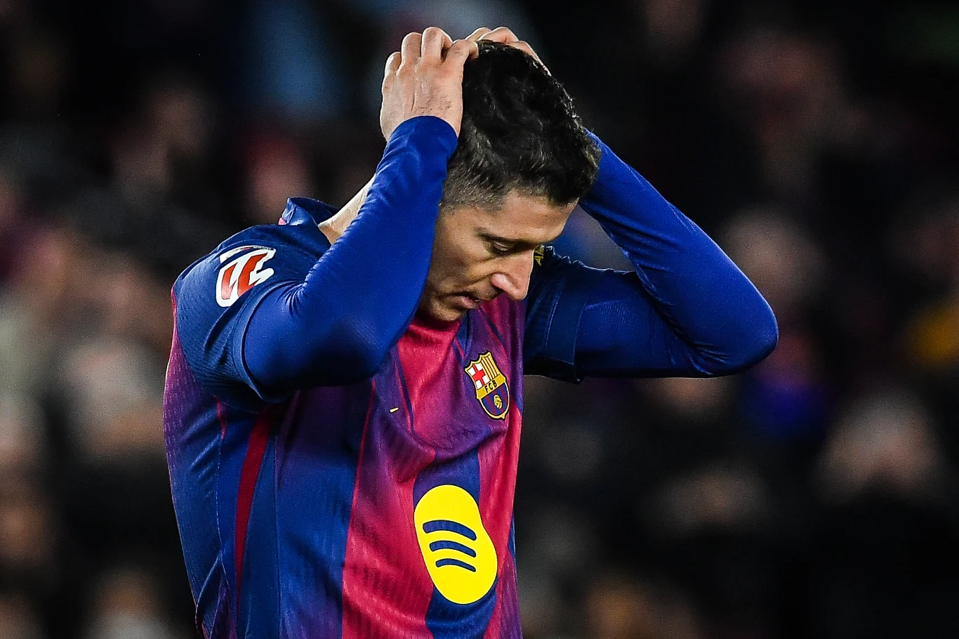 Zawodnik piłkarski w stroju FC Barcelona trzyma ręce na głowie, wyrażając rozczarowanie. Na koszulce widoczne logo klubu oraz sponsorów. Tło rozmyte, skupiające uwagę na postaci sportowca.