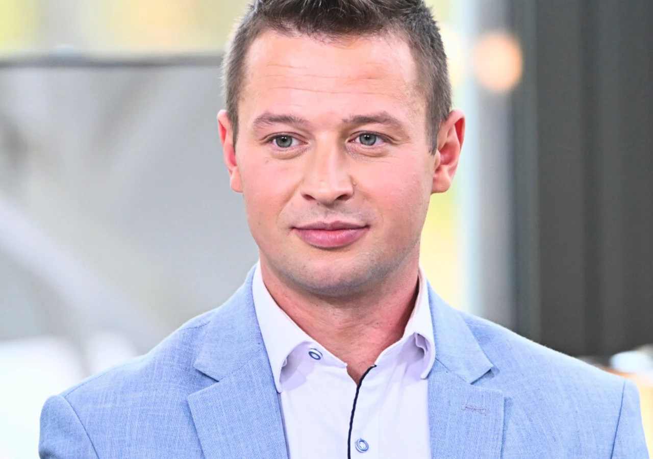 Krzysztof z "Rolnik szuka żony" 12 Mężczyzna w jasnoniebieskiej marynarce i białej koszuli, lekko uśmiechnięty, siedzi w dobrze oświetlonym wnętrzu.