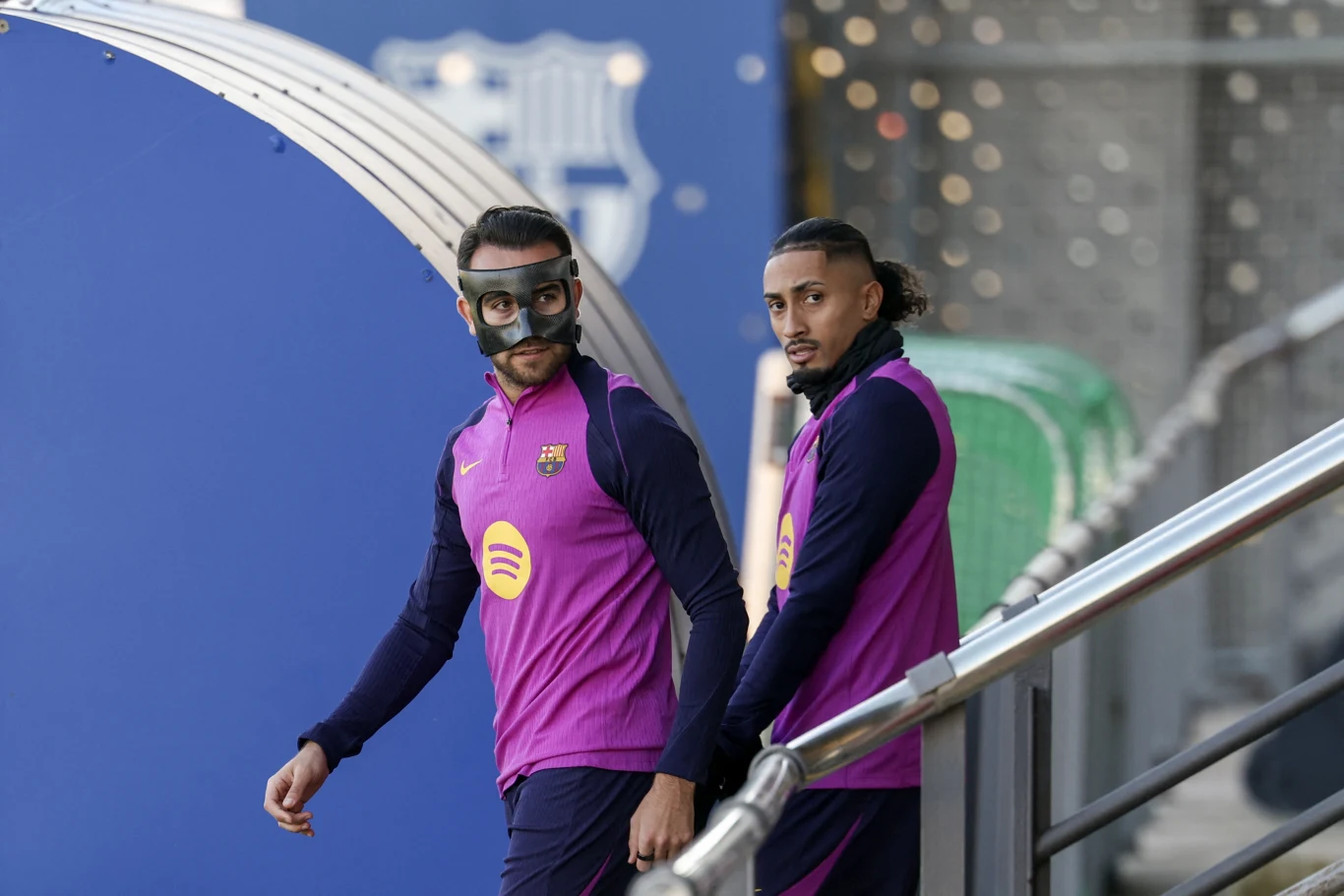 Dwóch piłkarzy ubranych w fioletowe stroje treningowe klubu FC Barcelona wychodzi z tunelu na stadion. Jeden z zawodników nosi ochronną maskę na twarzy.