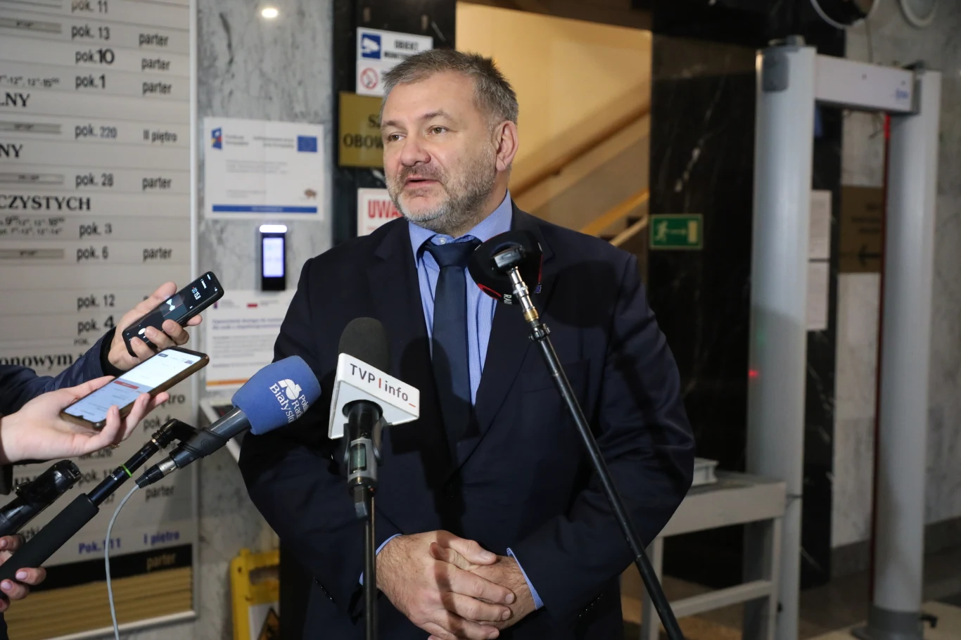 Waldemar Żurek na briefingu prasowym w Białymstoku. Zdjęcia ilustracyjne Mężczyzna w garniturze udziela wywiadu kilku dziennikarzom w holu budynku użyteczności publicznej, przed nim mikrofony różnych mediów, w tle tablica informacyjna i wejście obok wykrywacza metali.