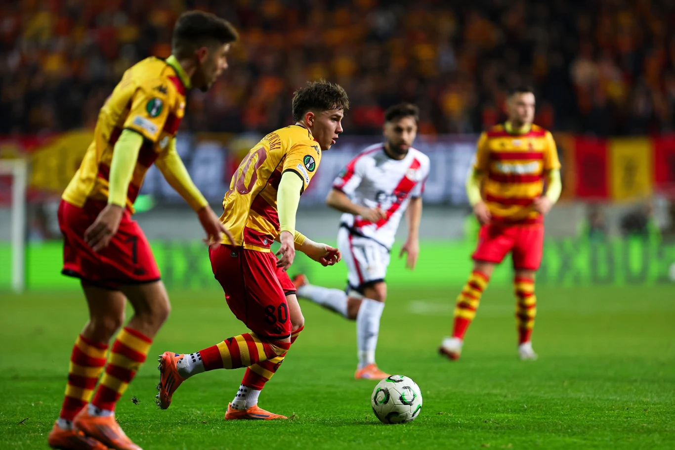 Oskar Pietuszewski przy piłce Zawodnicy dwóch drużyn piłkarskich rywalizują ze sobą na boisku, piłkarz w żółto-czerwonym stroju prowadzi piłkę, a pozostali uczestnicy są skoncentrowani na grze, w tle widoczne trybuny pełne kibiców.