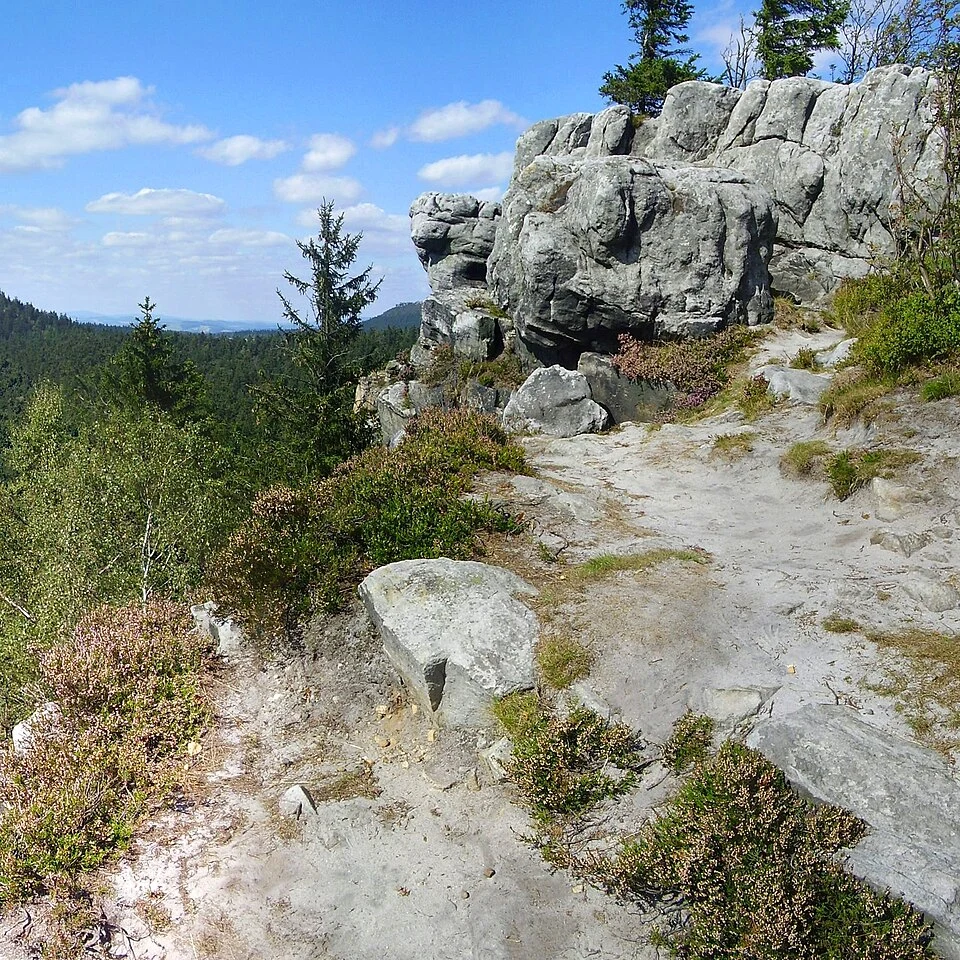 Narożnik skrywa mroczną tajemnicę Wyeksponowane skały na szczycie wzgórza otoczone gęstym lasem, z widokiem na zielone pagórki rozciągające się aż po horyzont, nad nimi rozproszone białe chmury na błękitnym niebie.