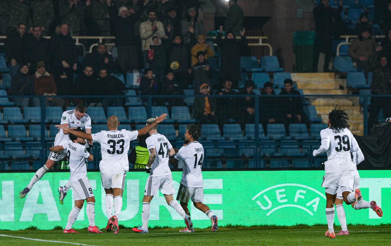 Grupa piłkarzy drużyny w białych strojach świętuje zdobycie gola na murawie stadionu piłkarskiego, w tle widoczni kibice na trybunach oraz neon z logotypem UEFA.
