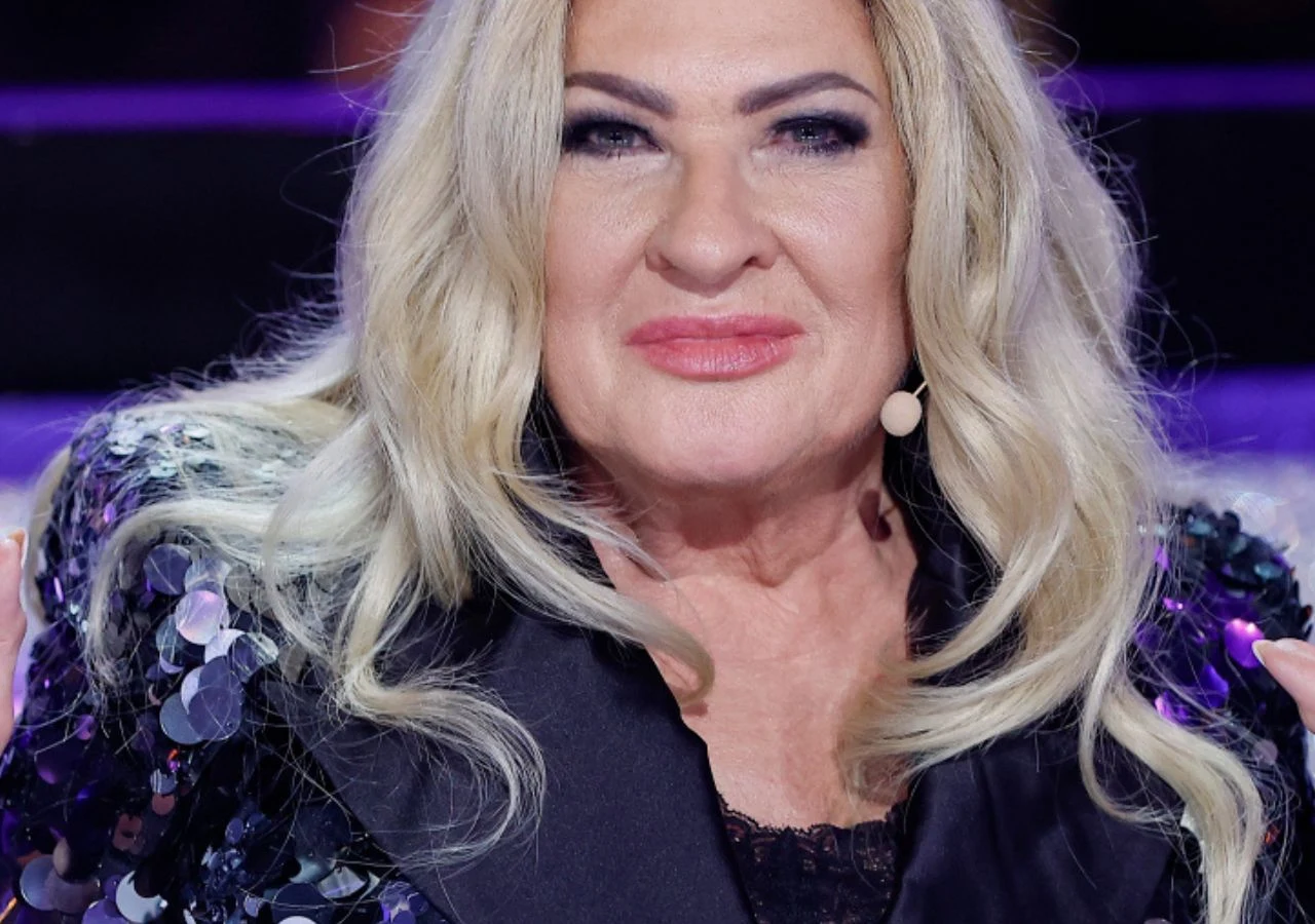 Kobieta w średnim wieku o blond włosach, uśmiechnięta, ubrana w cekinową marynarkę. Na twarzy pełny makijaż, w uchu mikrofon typu headset. Tło rozmyte, fioletowe światła sugerujące scenę rozrywkową.