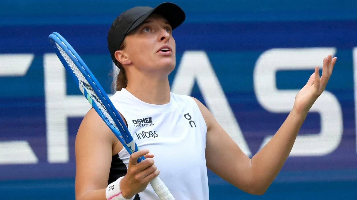 Zawodniczka tenisowa w białej koszulce z rakietą w ręku, patrząca do góry z wyrazem zdziwienia lub rozczarowania na twarzy, na tle niebieskiej tablicy z napisem.