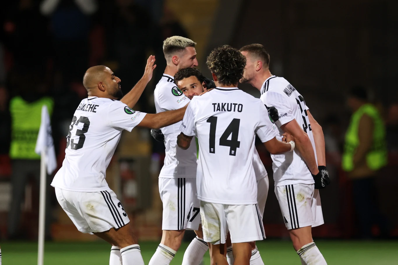 Grupa piłkarzy w białych strojach świętuje zdobycie gola na boisku, obejmując się i okazując radość z sukcesu sportowego, w tle widoczni są pozostali uczestnicy spotkania oraz fragment stadionu.