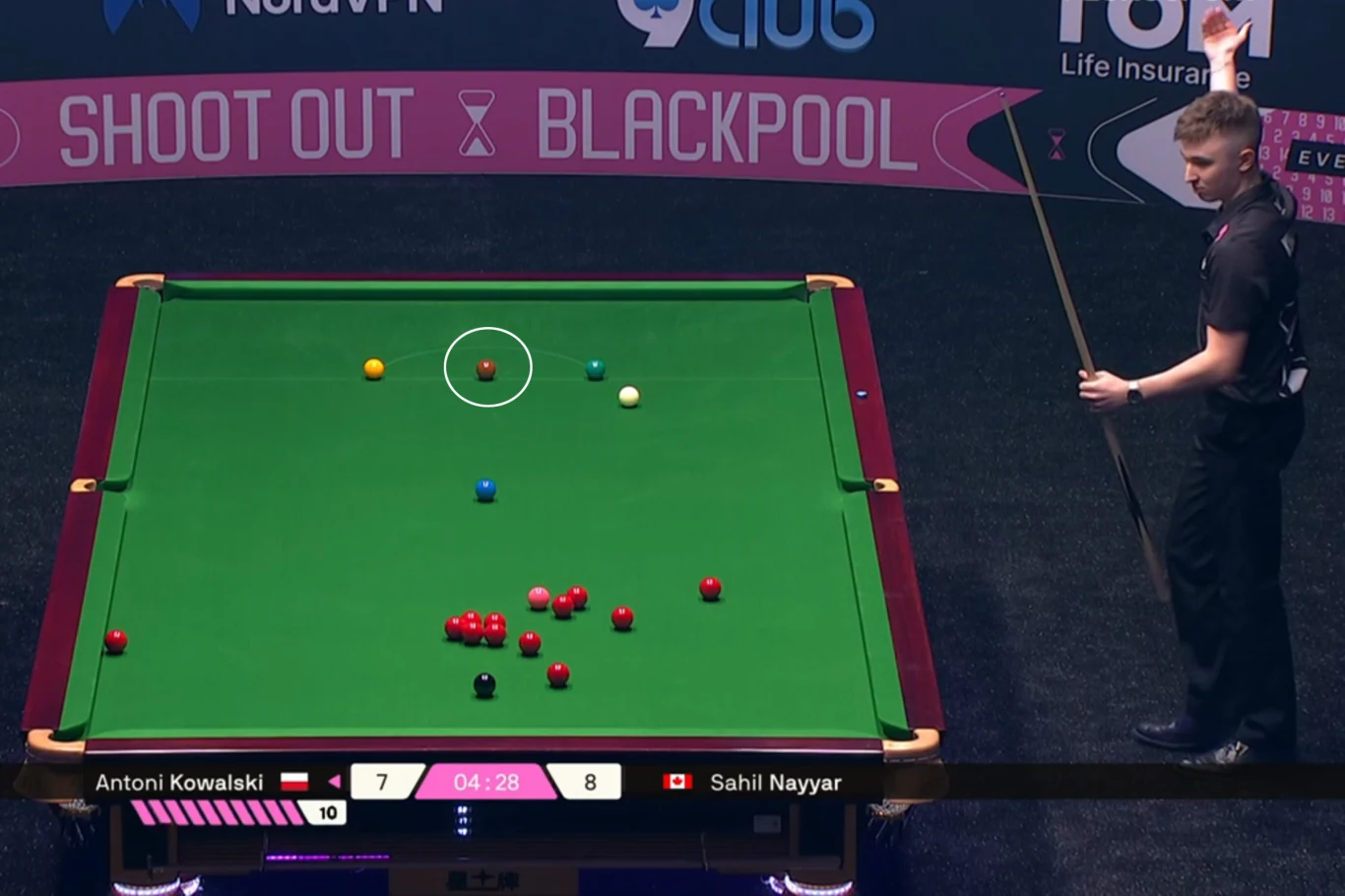 Stół bilardowy do snookera z rozłożonymi bilami, dwóch zawodników: jeden stoi przy stole z kijem, na ekranie widoczne są ich nazwiska i aktualny wynik meczu. W tle widoczne są reklamy i nazwa turnieju Shoot Out Blackpool.