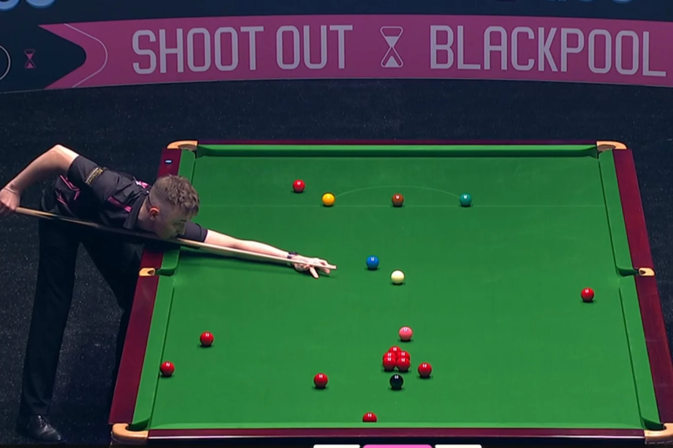 Zawodnik snookera wykonuje precyzyjny strzał na zielonym stole podczas turnieju, wokół rozmieszczone są kolorowe bile, a w tle widoczny jest baner z napisem Shoot Out Blackpool.