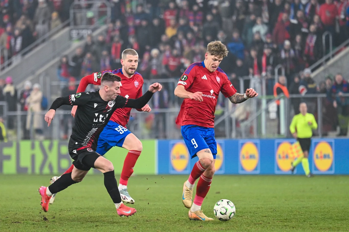 Grupa piłkarzy walcząca o piłkę na boisku podczas meczu, z wyraźnie widocznymi zawodnikami w czerwonych i czarnych strojach oraz tłumem kibiców oglądających spotkanie na trybunach.