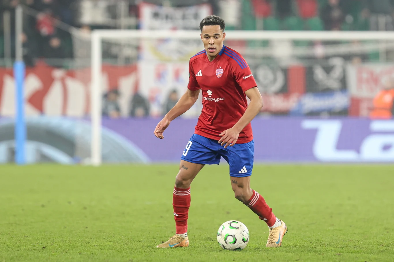 Piłkarz w czerwono-niebieskim stroju przygotowuje się do poprowadzenia piłki podczas meczu na murawie stadionu, w tle widać kibiców i transparenty.