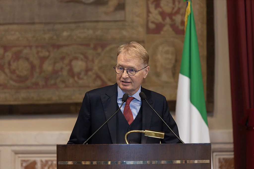 Włoski senator Lucio Malan Mężczyzna w średnim wieku w eleganckim garniturze i czerwonym krawacie przemawia z mównicy, w tle włoska flaga oraz ozdobny gobelin.