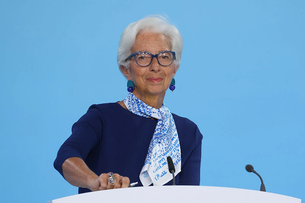 Szefowa Europejskiego Banku Centralnego Christine Lagarde Starsza kobieta z siwymi włosami i okularami, w eleganckim granatowym stroju z apaszką, stoi za mównicą na tle jasnoniebieskiej ściany, uśmiechając się delikatnie.