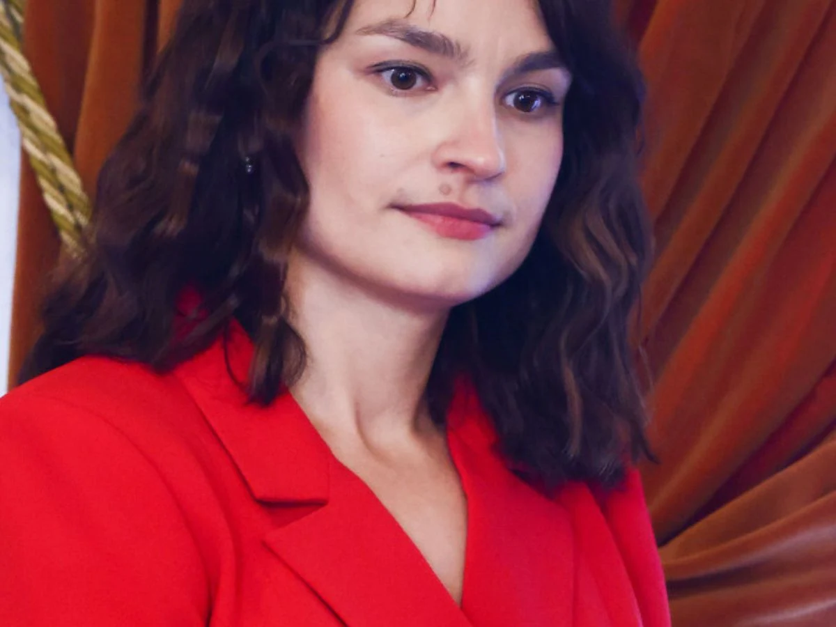 Anna Starmach Kobieta o ciemnych falowanych włosach, ubrana w czerwony żakiet, stoi na tle brązowej zasłony z ozdobnym sznurem po lewej stronie kadru.