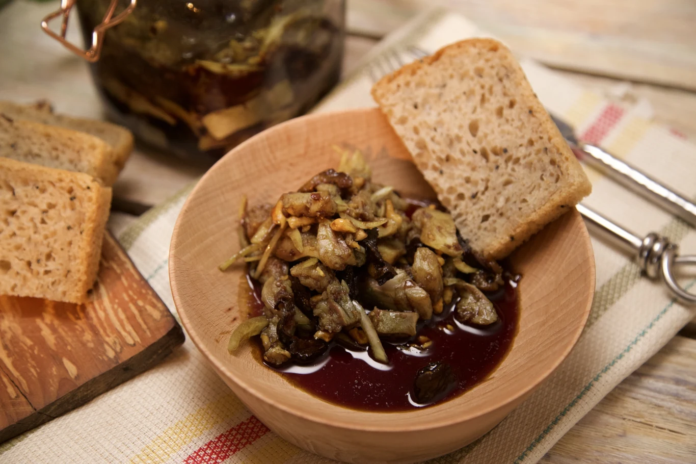 Śledzie w oleju dyniowym Drewniana miska wypełniona marynowanymi grzybami w sosie, obok kawałek chleba na krawędzi miski i kilka kromek chleba na desce w tle. Wszystko ułożone na jasnej serwetce z widelcem w tle.