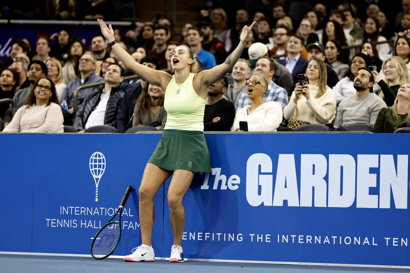 Tenisistka w jasnożółtym topie i zielonej spódniczce wyraża emocje na korcie podczas meczu, trzymając ręce w górze. W tle widoczna publiczność oraz plansza z napisem 'The Garden'.