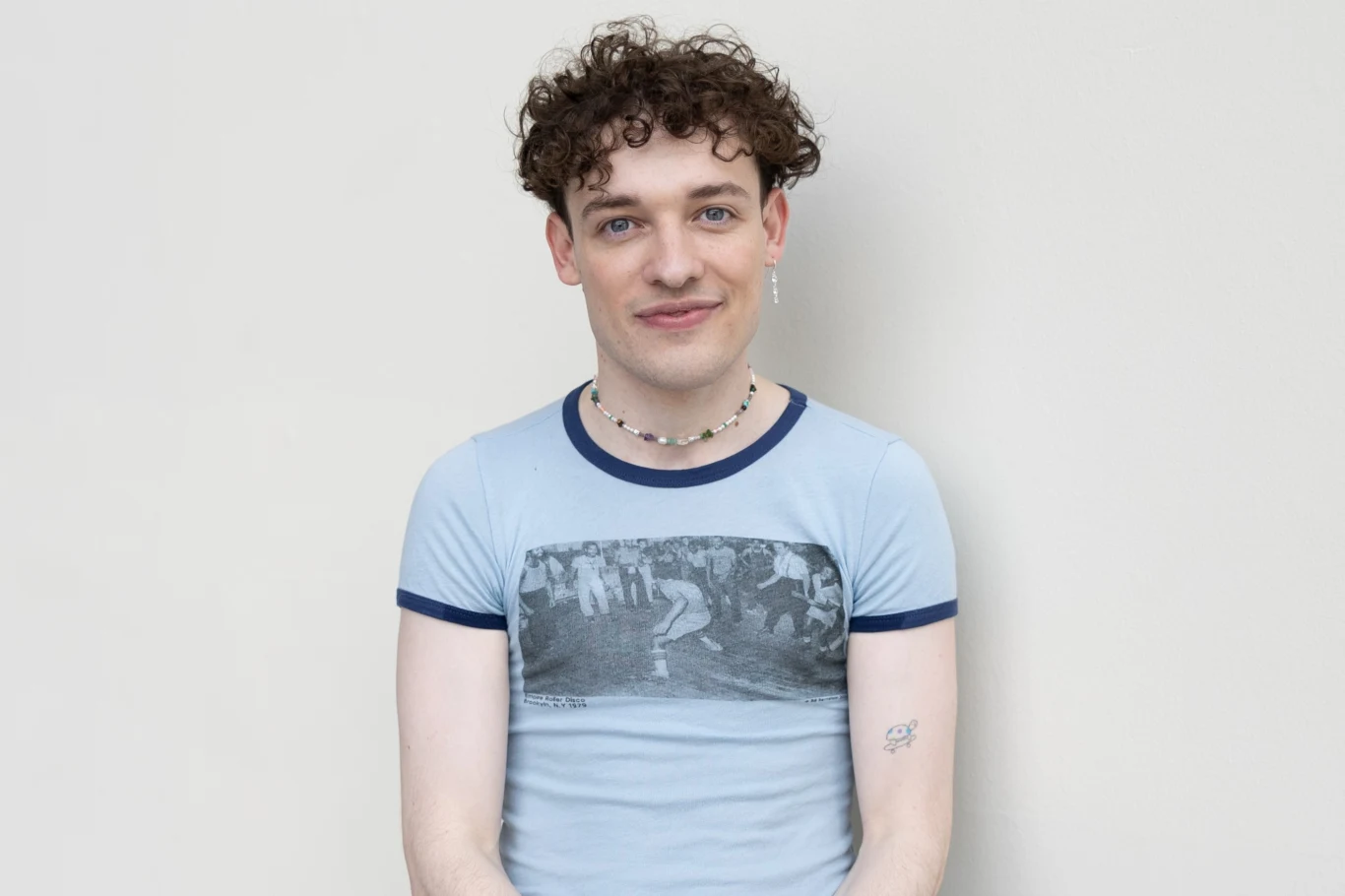 Nemo zwyciężyli podczas Eurowizji 2024 Młody mężczyzna z kręconymi włosami ubrany w niebieską koszulkę z grafiką i naszyjnikiem z koralików, z delikatnym uśmiechem, siedzi na tle jasnej, jednolitej ściany.