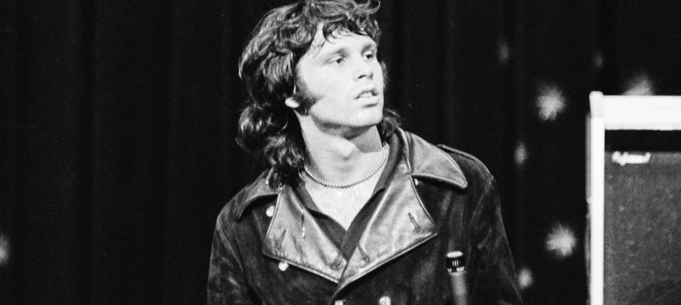 Jim Morrison dołączył do niesławnego "Klubu 27" Młody mężczyzna w skórzanej kurtce z długimi włosami patrzy w bok, w tle ciemne zasłony z jasnymi punktami i fragment wzmacniacza.