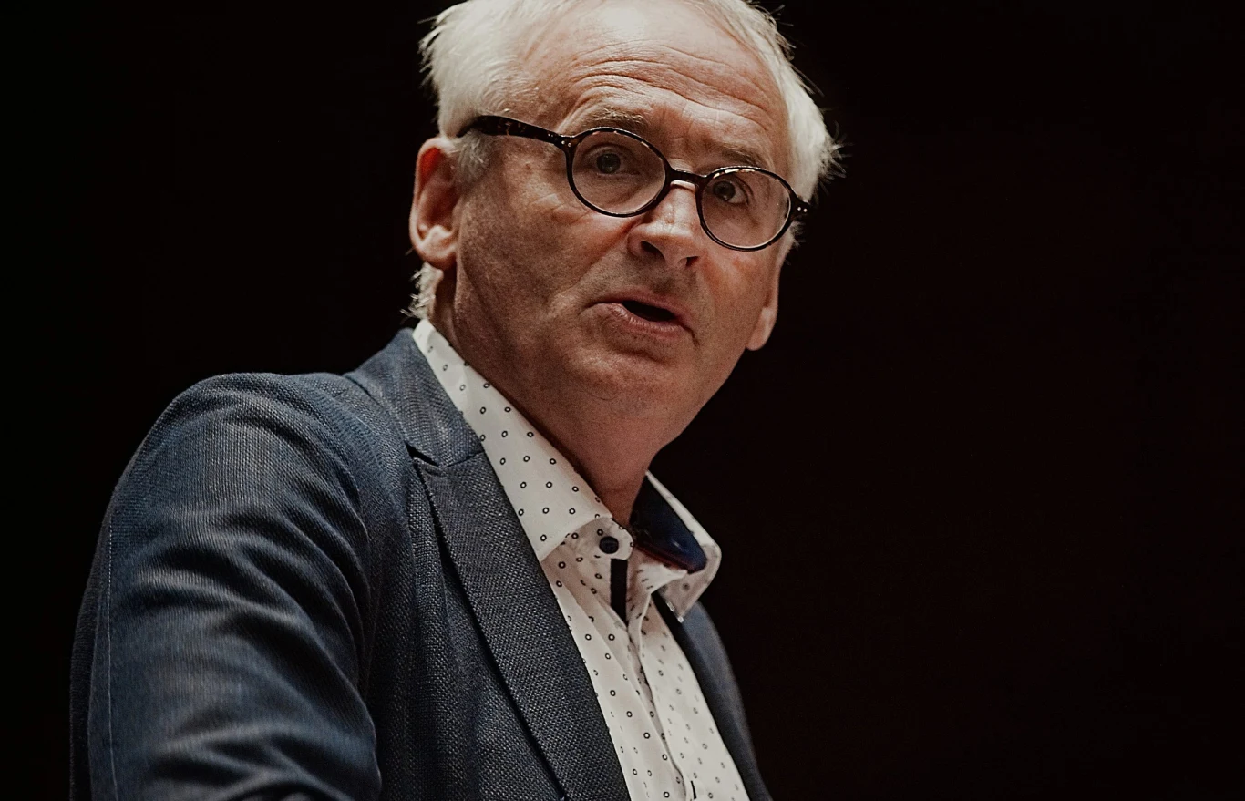 Krzesimir Dębski znów jest zakchany? Starszy mężczyzna w okularach i białych włosach ubrany w granatową marynarkę oraz koszulę w drobny wzór, patrzy w stronę obiektywu na ciemnym tle.