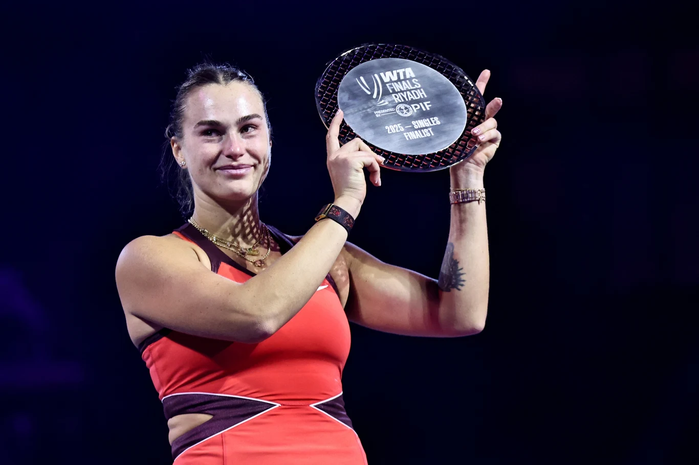Tenisistka w czerwonym stroju sportowym trzyma w rękach szklaną nagrodę z napisem WTA Finals Fort Worth, GPIF, pozując z uśmiechem na tle ciemnego, rozświetlonego światłem reflektorów tła.