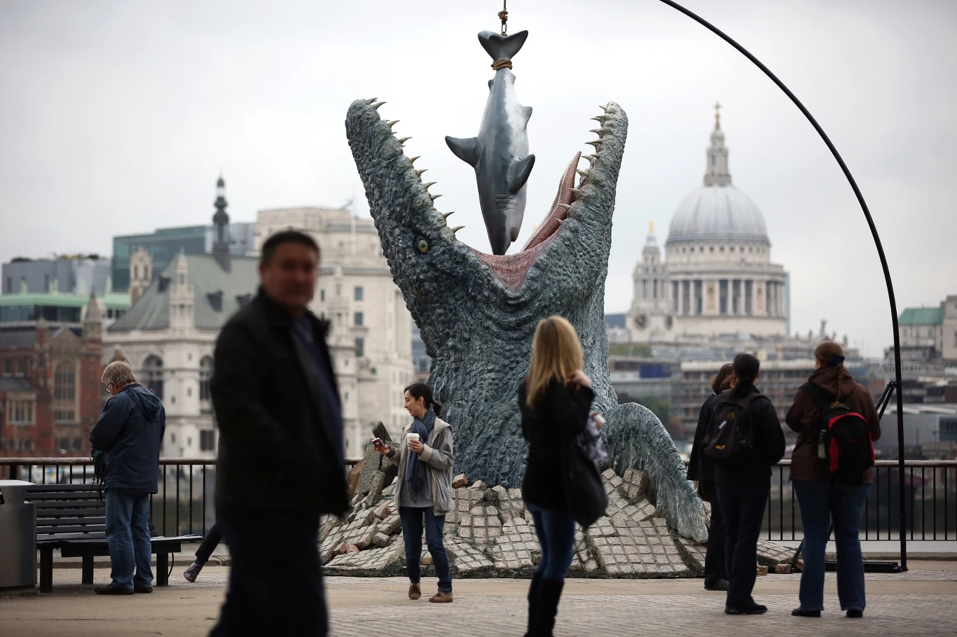 Ciekawy pomnik mozazaura z Londynu, nawiązujący do "Jurassic World" Monumentalna instalacja przedstawiająca otwartą paszczę prehistorycznego gada, który łapie zawieszonego na haku rekina, otoczona przez grupę spacerujących ludzi na tle miejskiej architektury.