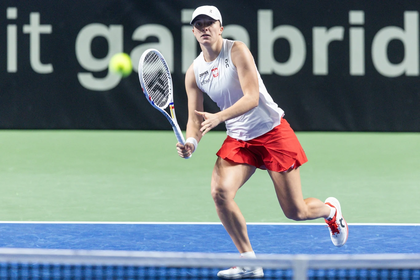 Tenisistka w białej czapce i stroju sportowym z rakietą w dłoni, skoncentrowana na odbiciu nadlatującej piłki na kortach twardych. W tle fragment bandy reklamowej.
