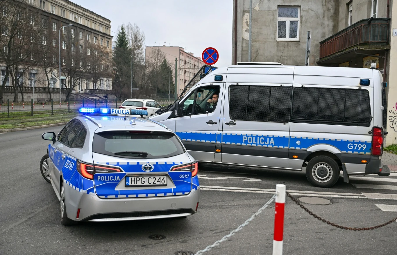 Radiowóz i policyjny furgon z włączonymi światłami, blokują wjazd na ulicę wśród miejskiej zabudowy; widoczne są znaki drogowe oraz fragmenty budynków.