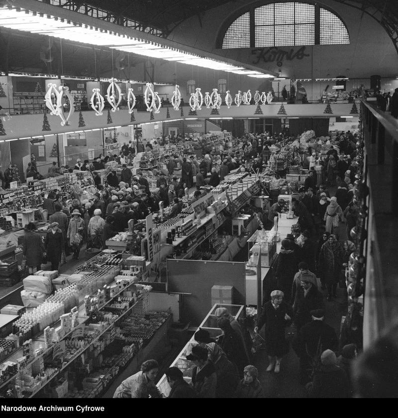 Tłum ludzi robiących zakupy w dużej, zadaszonej hali ozdobionej dekoracjami świątecznymi, rzędy stoisk z różnorodnym asortymentem, półki wypełnione towarami, atmosfera przygotowań do świąt.