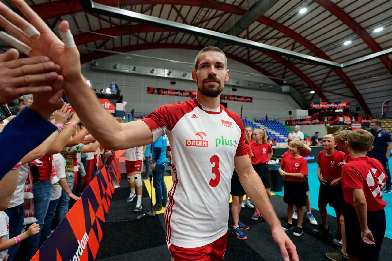 Siatkarz w stroju reprezentacji Polski z numerem 3 przybija piątki z kibicami w hali sportowej, w tle widoczna grupa dzieci i stojące osoby, trybuny częściowo zapełnione.