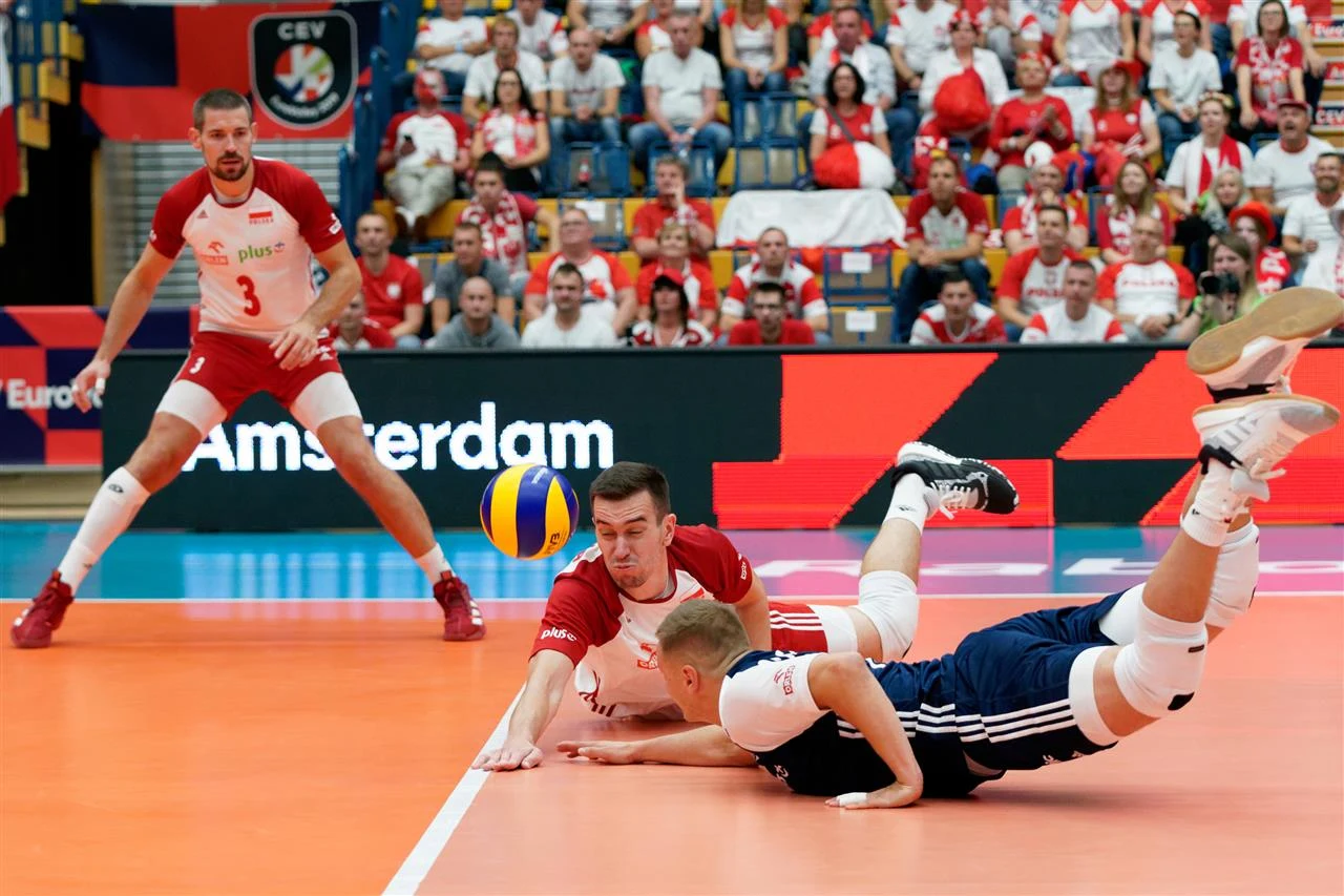 Dwóch siatkarzy rywalizuje o piłkę tuż nad podłogą boiska, jeden z nich leży na podłodze w poślizgu, drugi sięga po piłkę ręką, a w tle widać trzeciego zawodnika oraz liczną publiczność na trybunach.