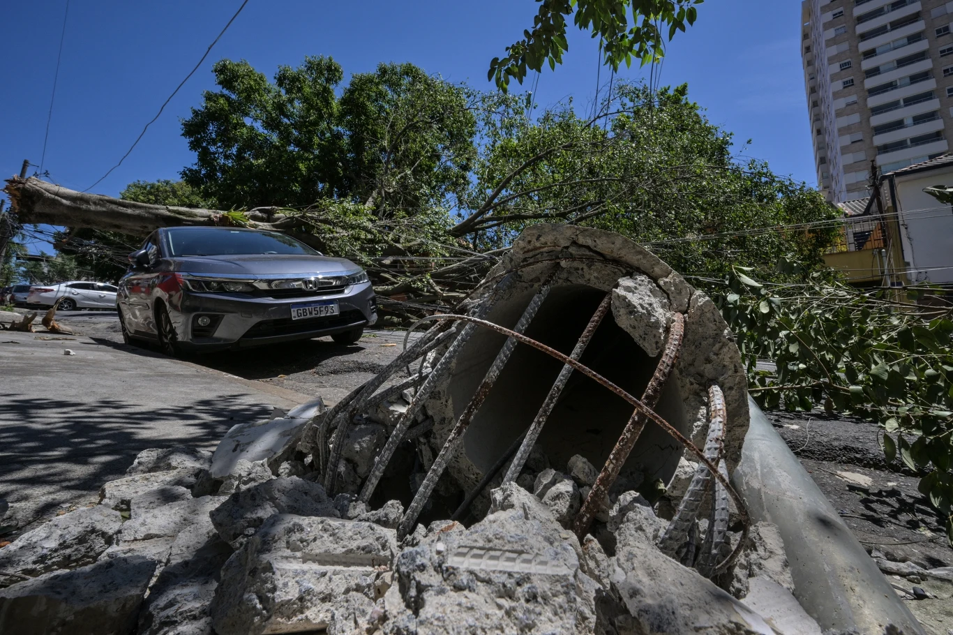 Skutki przejścia cyklonu przez Sao Paulo. Wichura powaliła w mieście 231 drzew Zniszczony słup betonowy oraz gałęzie przewróconych drzew leżą na ulicy obok zaparkowanego samochodu, w tle widać wysokie budynki i drzewa.