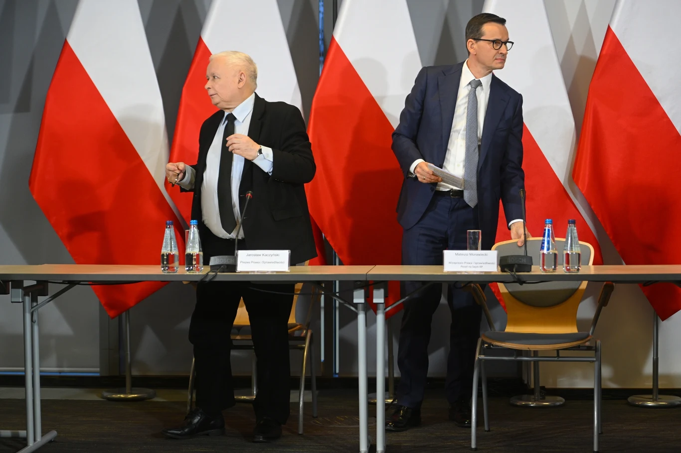 Jarosław Kaczyński i Mateusz Morawiecki Dwóch mężczyzn w garniturach stoi przy długim stole konferencyjnym, na którym ustawione są butelki wody i szklanki. W tle wywieszone są polskie flagi na tle geometrycznego wzoru. Jeden z mężczyzn trzyma dokumenty, obaj kierują wzrok w przeciwnych kieru...