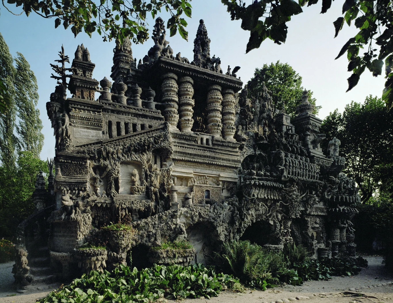 Monumentalna budowla z bogato zdobioną fasadą, pełna detali architektonicznych, wież i kolumn, otoczona zielenią drzew oraz krzewów w naturalnym otoczeniu.